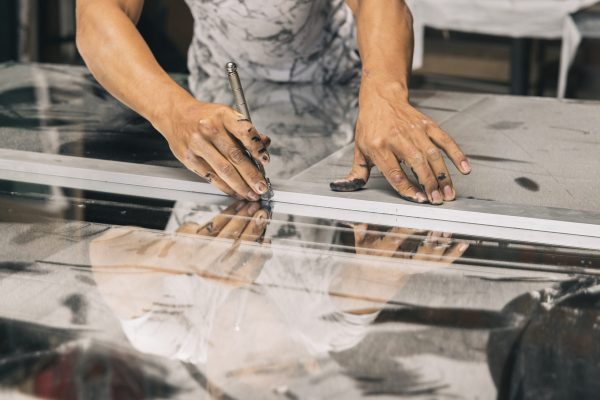 A close up of hands holding a glass cutter, scoring a piece of glass along measured lines in a workshop